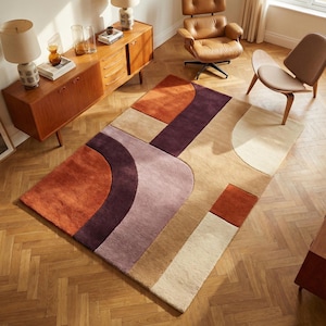Könnte beinhalten: Rechteckiger Teppich mit geometrischem Design in Orange-, Lila-, Beige- und Cremetönen. Der Teppich weist geschwungene und rechteckige Formen auf, die ein modernes, abstraktes Muster bilden. Der Teppich liegt auf einem Holzboden.