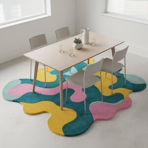 K&ouml;nnte beinhalten: Ein modernes Esszimmer-Setup mit einem hellen Holztisch und vier wei&szlig;en St&uuml;hlen. Unter dem Tisch liegt ein farbenfroher, abstrakter Teppich in T&uuml;rkis, Gelb und Rosa. Eine wei&szlig;e Kanne, eine Pflanze und Gl&auml;ser stehen auf dem Tisch.