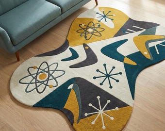 May include: An irregularly shaped area rug featuring a cream background with mustard yellow, teal, and dark gray geometric patterns. The design includes atomic starburst motifs, boomerang shapes, and orbital ring patterns across the surface.