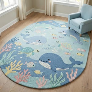 Puede incluir: Una alfombra ovalada con temática oceánica. Presenta ballenas, delfines, tortugas y peces de dibujos animados en tonos azules, verdes, amarillos y rosas. También incluye diseños de coral y algas marinas. La alfombra está sobre un suelo de madera.