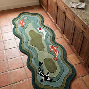 May include: An elongated, irregularly shaped area rug featuring a layered, wavy design in shades of green. The rug depicts four koi fish in orange, white, and black, swimming across the textured surface, which mimics the appearance of pond water.