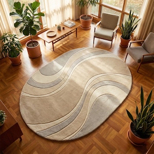 May include: An oval area rug with a wave design in shades of beige and gray. The rug is placed on a hardwood floor in a room with natural light and potted plants. The rug's dimensions are not visible.