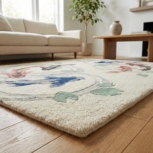 May include: Cream-colored area rug with a koi fish design. The rug features a repeating pattern of koi fish in shades of blue, pink, and gray, with green leaf accents. The rug is placed on a hardwood floor.
