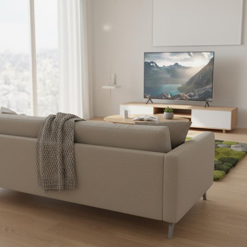 May include: A modern living room featuring a beige sofa with a gray throw blanket. A large flat-screen TV displays a mountain landscape. The room includes a wooden coffee table, a white and wood TV stand, and a green rug.
