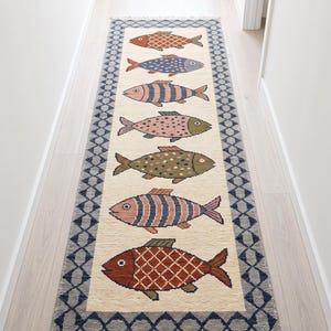 May include: A long rectangular runner rug with a cream-colored center featuring seven distinct fish illustrations in various colors and patterns. The rug is framed by a dark blue and gray diamond-shaped border and finished with fringe at both ends.