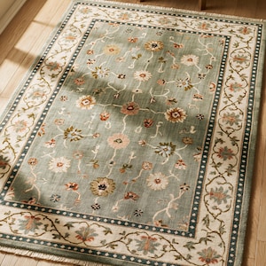 May include: A rectangular area rug featuring a sage green field with a floral pattern in shades of pink, cream, and brown. The rug has a wide cream border with a dark blue dotted line and a floral vine motif, finished with short white fringe.
