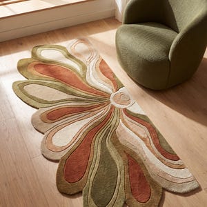 May include: A decorative rug with a floral design in shades of green, orange, and beige. The rug is placed on a wooden floor, near a window and a green armchair. The design features layered petal-like shapes radiating from a central point.