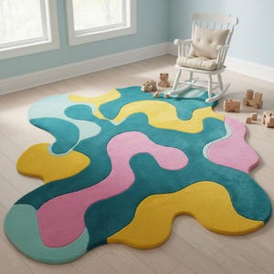 K&ouml;nnte beinhalten: Ein farbenfroher Teppich mit abstraktem Design. Er zeigt flie&szlig;ende Formen in T&uuml;rkis, Gelb, Rosa und Hellblau. Ein wei&szlig;er Schaukelstuhl und Holzspielzeug im Hintergrund deuten auf ein Kinderzimmer oder Spielzimmer hin.