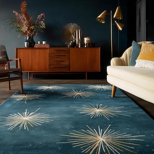 May include: A teal rug featuring a starburst design in white and gold. The rug is in a living room with a mid-century wooden cabinet, a white sofa, and a gold floor lamp. The room has a dark blue wall, creating a stylish interior.