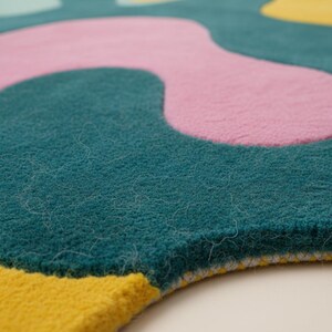 K&ouml;nnte beinhalten: Nahaufnahme eines farbenfrohen Teppichs mit abstraktem Design. Der Teppich hat einen tealfarbenen Hintergrund mit gro&szlig;en, unregelm&auml;&szlig;igen Formen in Rosa und Gelb. Die Textur des Teppichs wirkt weich und pl&uuml;schig, mit sichtbaren Fasern.