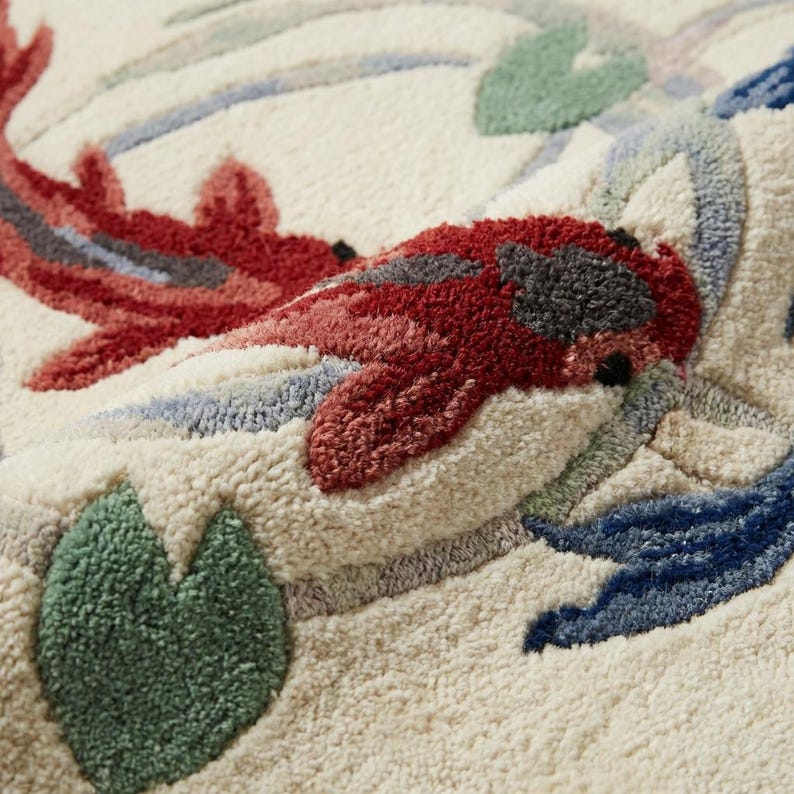May include: Close-up of a cream-colored rug with a koi fish design. The rug features two red and pink koi fish, green lily pads, and blue and gray water elements. The design is textured and detailed.