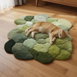 Op de afbeelding: Een groene, wolk-vormige vloerkleed met verschillende groentinten. Een slapende oranje tabby kat ligt op het kleed. Het kleed is gemaakt van een zacht, getextureerd materiaal en ligt op een houten vloer.