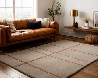 Puede incluir: Una alfombra rectangular beige con un diseño de cuadrícula de líneas naranjas. Se encuentra sobre un suelo de madera en un salón, con un sofá de cuero marrón y otros muebles en el fondo. Dimensiones aproximadas: 200 cm x 300 cm.