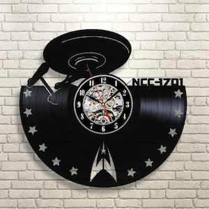 May include: A black vinyl record clock featuring a Star Trek Enterprise spaceship design. The clock face has a silver rim with black Roman numeral markings and a silver center with a clock mechanism. The clock is decorated with silver stars and the text "NCC-1701".