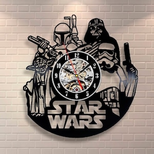 Puede incluir: Reloj de pared negro con temática de Star Wars con Darth Vader, Yoda, Boba Fett y un Stormtrooper. El reloj tiene una esfera negra con números y manecillas blancas. Las palabras "STAR WARS" están en la parte inferior.