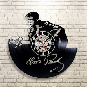 May include: Black vinyl record clock featuring a silhouette of Elvis Presley playing a guitar. The clock face has Roman numerals and a silver signature that reads "Elvis Presley".