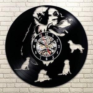 Puede incluir: Un reloj de vinilo negro con una silueta de perro y recortes de perros. La esfera del reloj tiene un movimiento mecánico visible con números en blanco y negro. Las manecillas del reloj son rojas y negras.