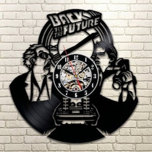 Può includere: Orologio in vinile nero con una silhouette di personaggi del film Ritorno al futuro. Il quadrante dell'orologio è rotondo, color argento con numeri e lancette neri. L'orologio è posizionato su uno sfondo di muro di mattoni.