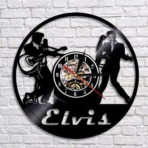 May include: Black vinyl record clock featuring a silhouette of Elvis Presley singing and playing a guitar. The clock face is a black circle with white numbers and a gold clock mechanism. The clock is titled "Elvis".