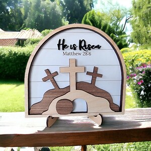 May include: Wooden Easter sign with a white background and a brown arch. The sign says "He is Risen" with the scripture reference "Matthew 28:6". The sign also features three wooden crosses and a stone tomb.