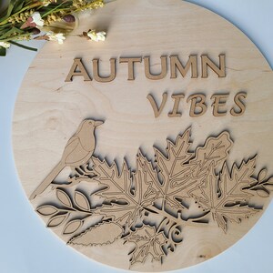 May include: A round wooden sign with the text "AUTUMN VIBES" in brown lettering. A bird and leaves are cut out of the wood and are also brown.
