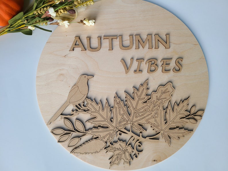 May include: A round wooden sign with the text "AUTUMN VIBES" in brown lettering. A bird and leaves are cut out of the wood and are also brown.