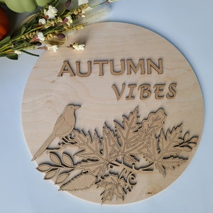 May include: A round wooden sign with the text "AUTUMN VIBES" in brown letters. The sign features a cutout design of a bird perched on a branch with leaves.