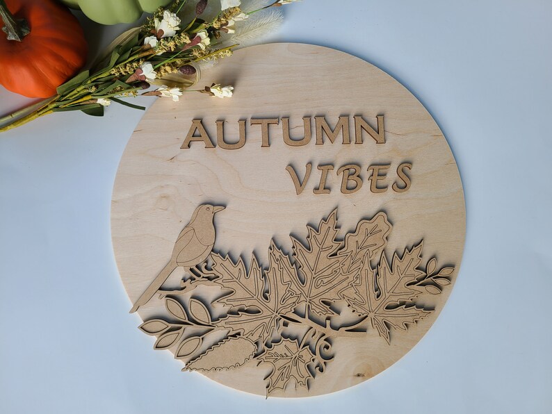 May include: A round wooden sign with the text "AUTUMN VIBES" in brown letters. The sign features a cutout design of a bird perched on a branch with leaves.