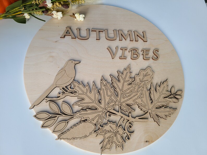 May include: A round wooden sign with the text "AUTUMN VIBES" in cutout letters. A bird is perched on a branch with leaves.