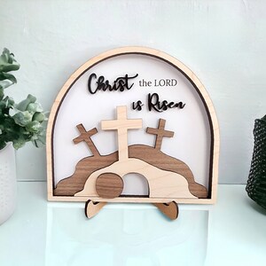 May include: Wooden Easter sign with a white background and the text "Christ the LORD is Risen". The sign features a 3D design of a tomb with a rolling stone, three crosses, and a hill.
