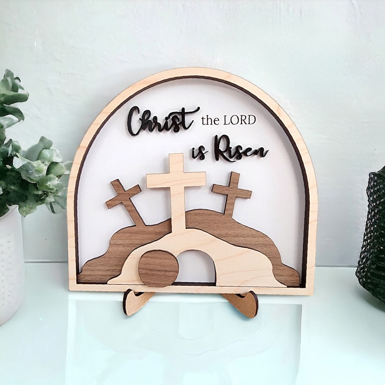 May include: Wooden Easter sign with a white background and the text "Christ the LORD is Risen". The sign features a 3D design of a tomb with a rolling stone, three crosses, and a hill.