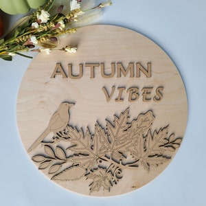 May include: A round wooden sign with the words "Autumn Vibes" in brown lettering. The sign features a stylized bird perched on a branch with leaves.