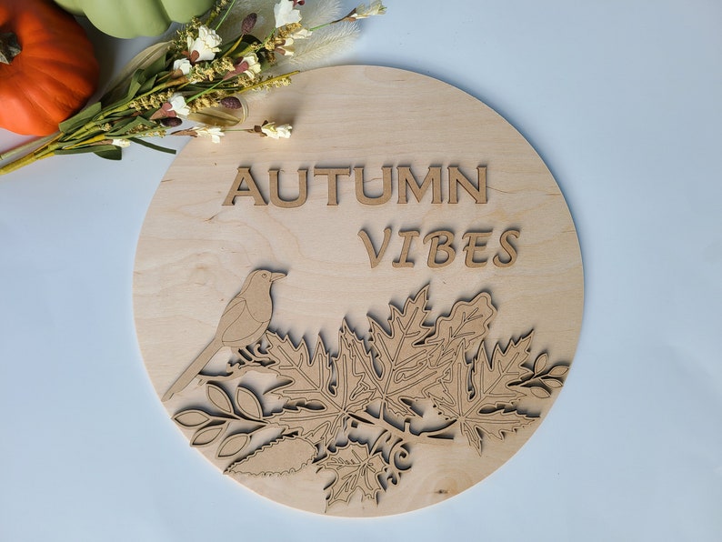 May include: A round wooden sign with the words "Autumn Vibes" in brown lettering. The sign features a stylized bird perched on a branch with leaves.