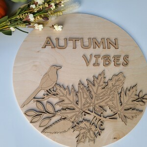 May include: A round wooden sign with the words "Autumn Vibes" in cutout letters. The sign features a cutout design of a bird perched on a branch with leaves.