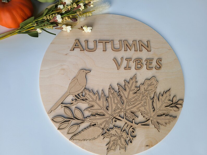 May include: A round wooden sign with the words "Autumn Vibes" in cutout letters. The sign features a cutout design of a bird perched on a branch with leaves.