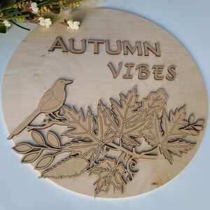 May include: A wooden round sign with the text "AUTUMN VIBES" in brown lettering. The sign features a cutout design of a bird perched on a branch with leaves.