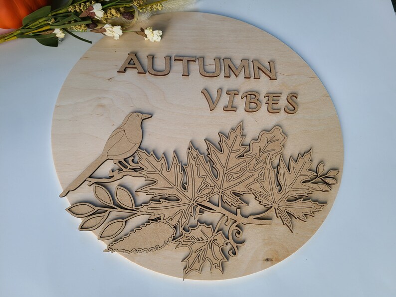 May include: A wooden round sign with the text "AUTUMN VIBES" in brown lettering. The sign features a cutout design of a bird perched on a branch with leaves.