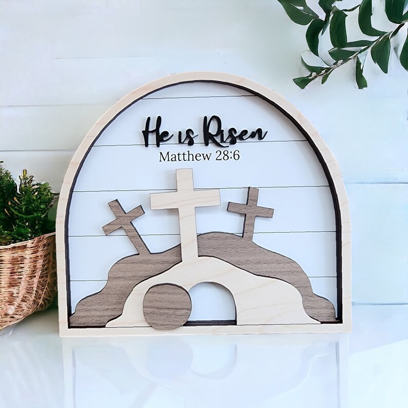 May include: Wooden Easter sign with a white background and a brown wooden arch. The sign says "He is Risen" with the scripture reference "Matthew 28:6". The sign also features three wooden crosses and a stone rolling away from a tomb.