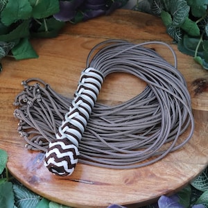 May include: A brown and white braided accessory with a rounded end and a bundle of grey cords. The accessory is displayed on a wooden surface, surrounded by green foliage.