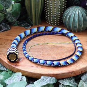 May include: A decorative whip with a braided design in shades of blue, yellow, and white. The whip is coiled on a wooden surface, with a round, patterned handle. The background includes green plants and decorative ceramic spheres.