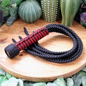 May include: A black and red braided whip with a braided handle. The whip is coiled on a wooden surface.