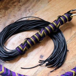 Puede incluir: Un látigo negro con un mango trenzado con detalles morados, negros y dorados. El mango del látigo tiene un anillo de metal en la parte superior. El látigo está sobre una superficie de madera.