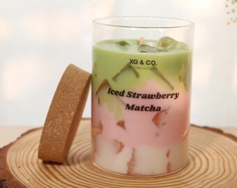May include: A layered candle in a clear glass, resembling an iced drink, with green, pink, and white sections. The candle has the text "Iced Strawberry Matcha". A cork coaster sits next to the candle on a wooden slice.