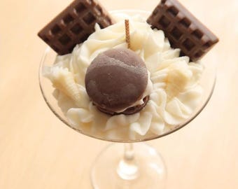 May include: A dessert sundae in a clear glass. The sundae features whipped cream, two chocolate waffles, and a chocolate macaron. A small wooden stick is placed in the centre of the dessert.