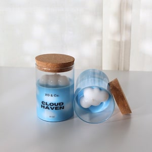 Puede incluir: Dos velas en tarro de cristal con tapas de corcho. Las velas tienen una base azul con un diseño de cera en forma de nube blanca. El texto "XG & Co." y "CLOUD HAVEN" está impreso en el tarro. Una vela está en posición vertical y la otra está de lado.