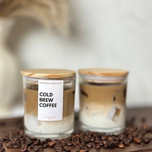 May include: Two glass candle jars with wooden lids. The jars are filled with a layered candle that resembles iced coffee. The candles are on a wooden surface with coffee beans scattered around them. The label on one jar reads "Cold Brew Coffee" and "The Copper House 11.5 oz".