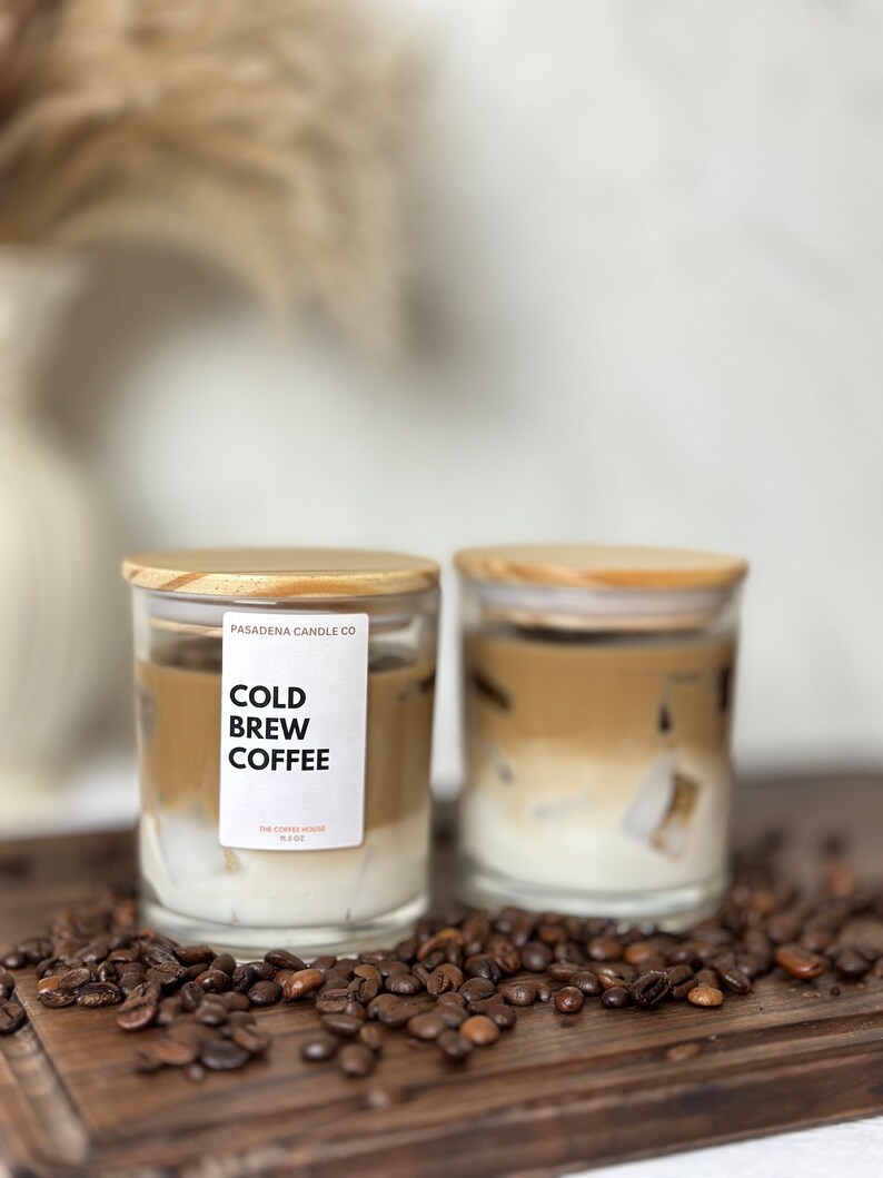 May include: Two glass candle jars with wooden lids. The jars are filled with a layered candle that resembles iced coffee. The candles are on a wooden surface with coffee beans scattered around them. The label on one jar reads "Cold Brew Coffee" and "The Copper House 11.5 oz".
