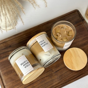 May include: Three glass candle jars with wooden lids. The candles are labelled "Cold Brew Coffee" and have a brown and white layered design. The candles are on a wooden surface.