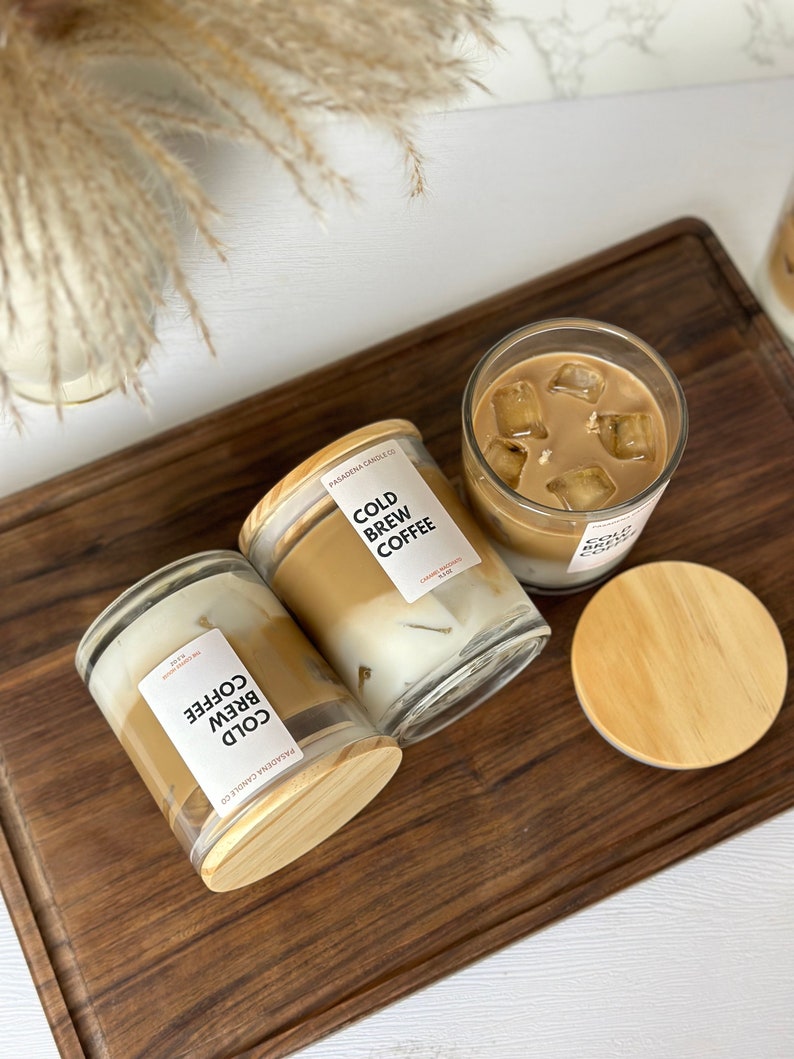May include: Three glass candle jars with wooden lids. The candles are labelled "Cold Brew Coffee" and have a brown and white layered design. The candles are on a wooden surface.