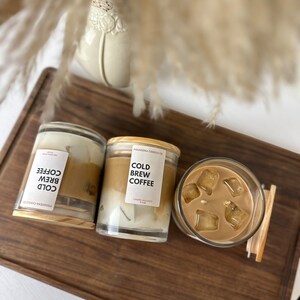 May include: Two glass candle jars with wooden lids and a glass of iced coffee on a wooden surface. The candles are labelled "Cold Brew Coffee" and the coffee is in a clear glass with ice cubes.